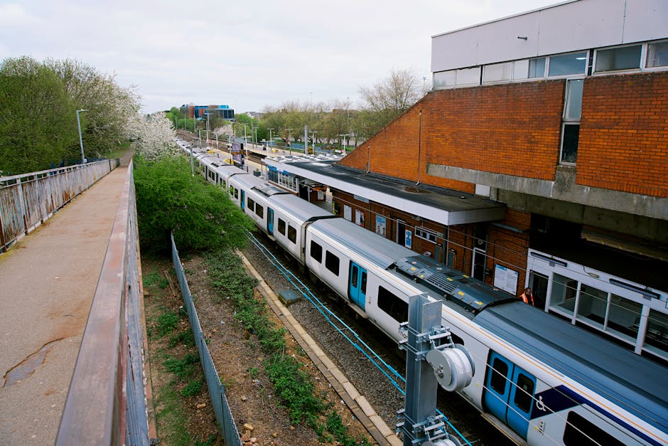 A railway station platform with a modern train stopped on the tracks, viewed from an elevated walkway on the left side of the image. The platform features a brick building with windows and signage, al