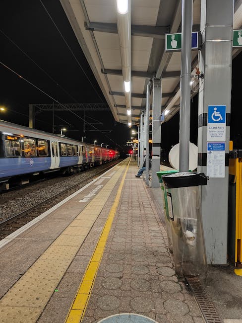 A railway station platform with a modern train stopped on the tracks, viewed from an elevated walkway on the left side of the image. The platform features a brick building with windows and signage, al