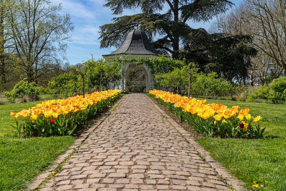 A scenic garden scene featuring a straight cobblestone pathway leading to a decorative white gazebo with a black dome-shaped roof, surrounded by lush green trees and shrubs. Flanking the pathway are v