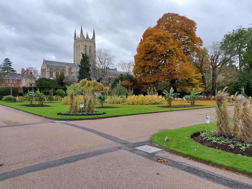 A scenic garden scene featuring a straight cobblestone pathway leading to a decorative white gazebo with a black dome-shaped roof, surrounded by lush green trees and shrubs. Flanking the pathway are v